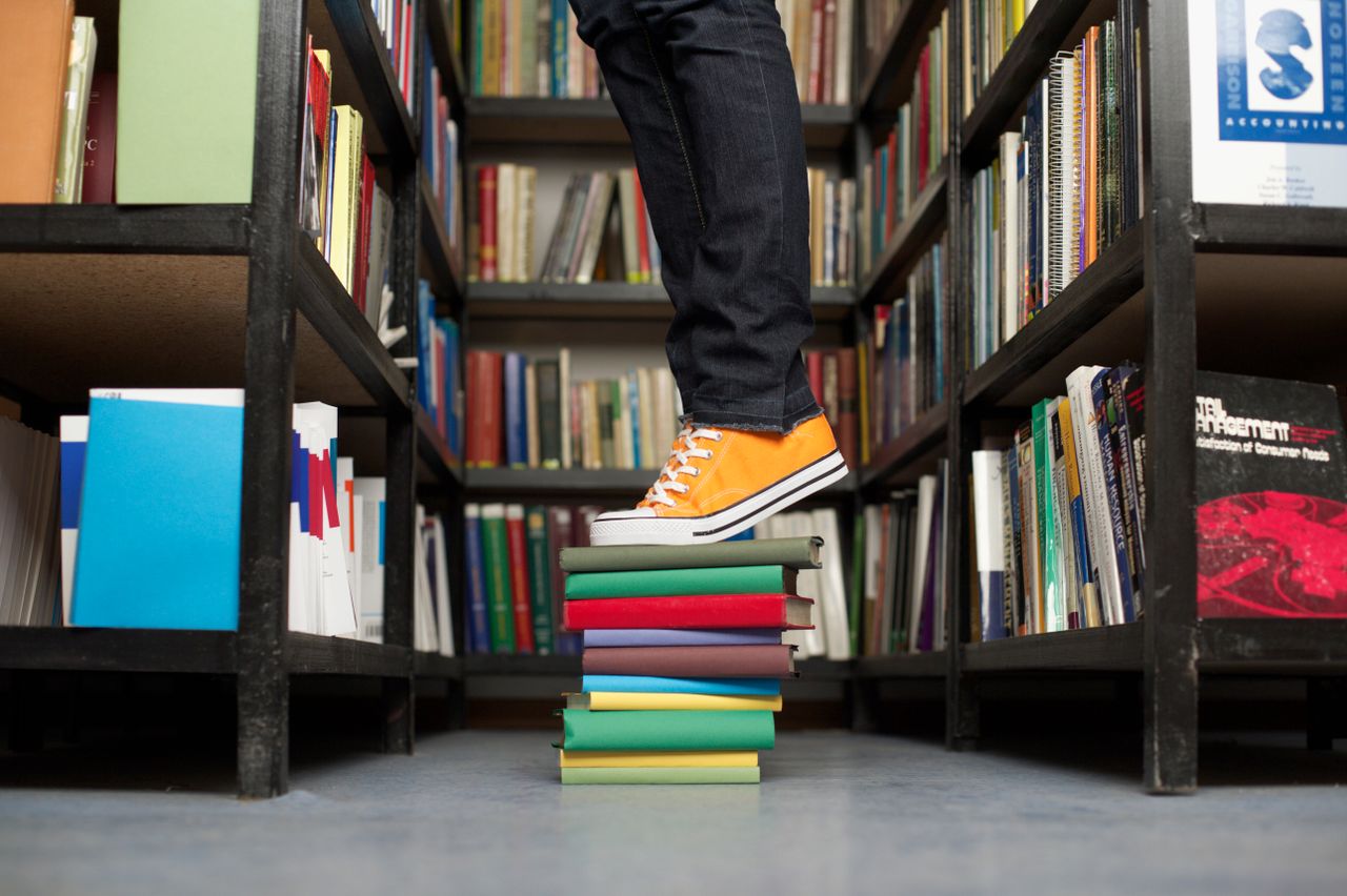 A person standing on a colorful stack of books in a library aisle.
