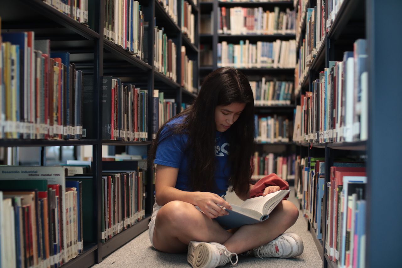 A young woman sitting on the floor of a library, reading a book intently.