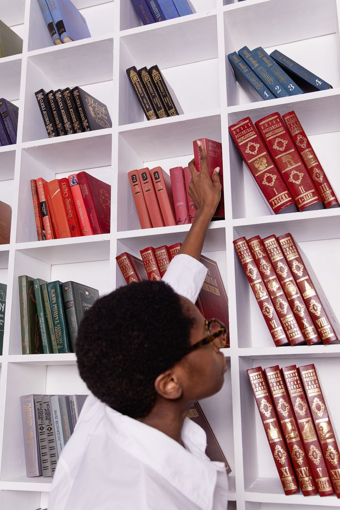 A stylish black woman reaches for a book on colorful shelves, embodying elegance and knowledge.